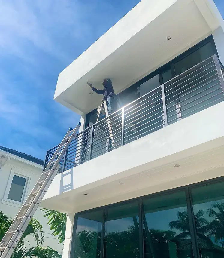 A Creative Images painter on an extension ladder brushing the underside of a white cantilevered soffit against a deep blue South Florida sky