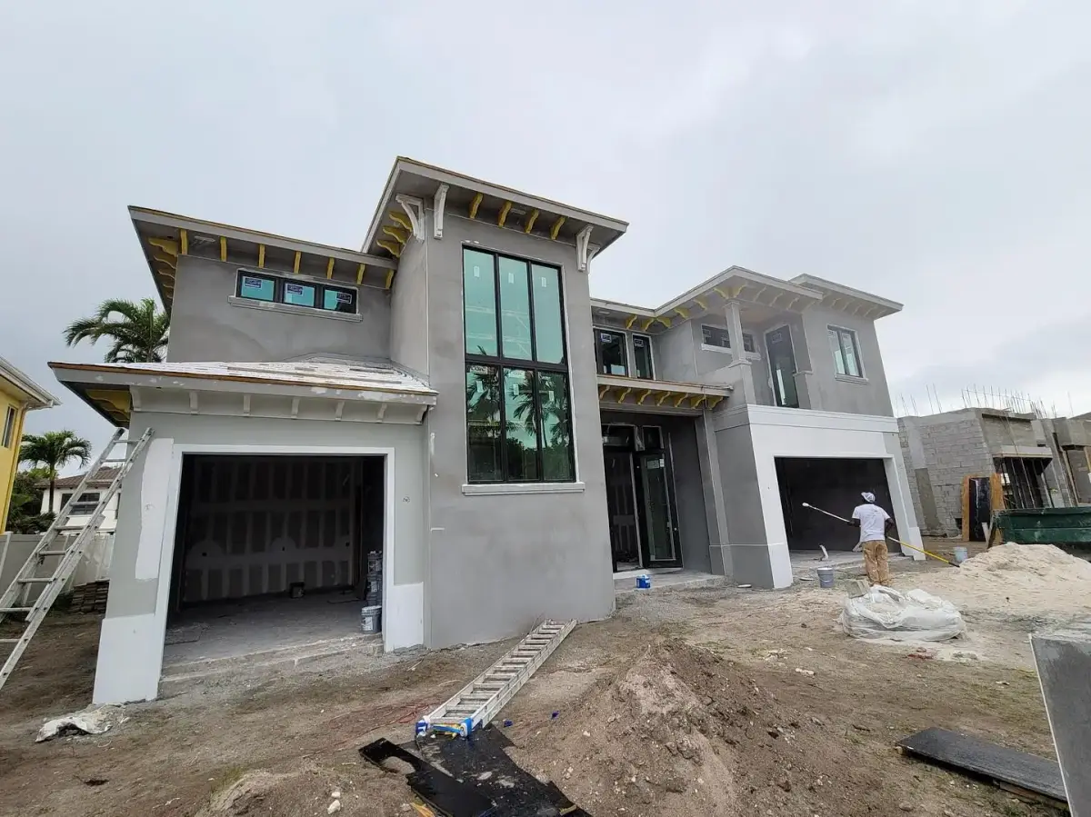 A painter spraying stucco on an active new-construction house with sheathing, ladders and neighbouring builds visible