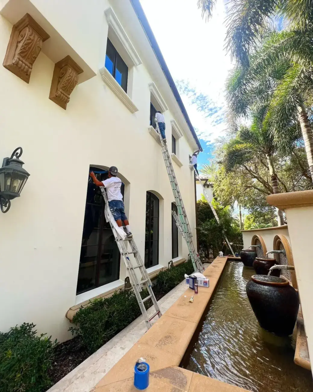 Two Creative Images painters on extension ladders working the exterior wall and window trim of a luxury Boca Raton home beside a water feature