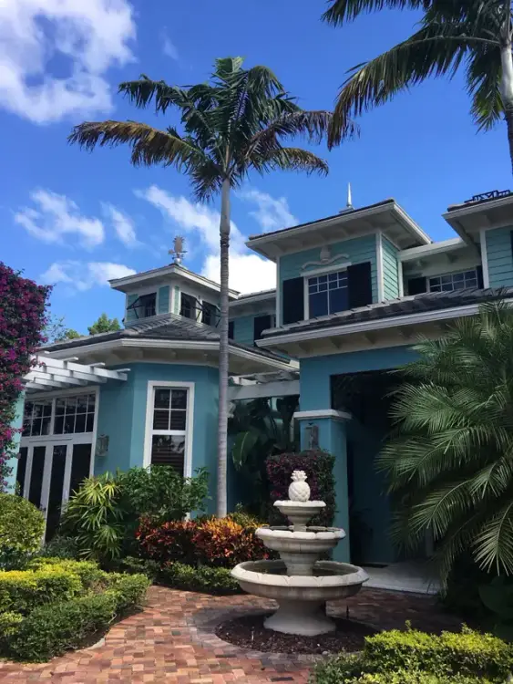 A turquoise Key West style home with white trim, a cupola and a three-tier stone pineapple fountain, painted by Creative Images Painting